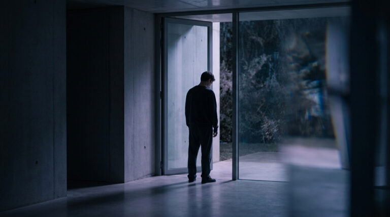 Un homme, de dos, se tient devant une porte vitrée ouverte, regardant vers l'extérieur sombre. Scène d'introspection.