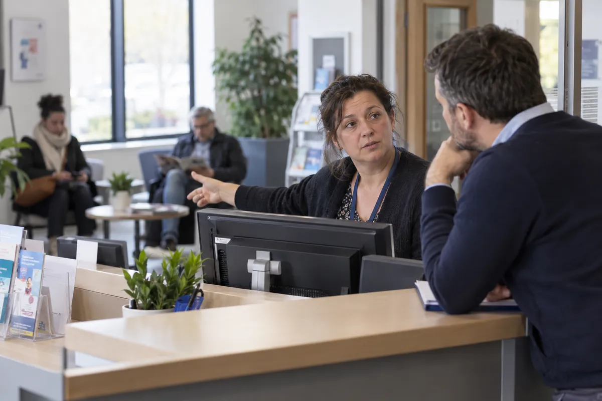 Deux collègues échangent dans un espace d’accueil avec un comptoir et une zone d’attente.