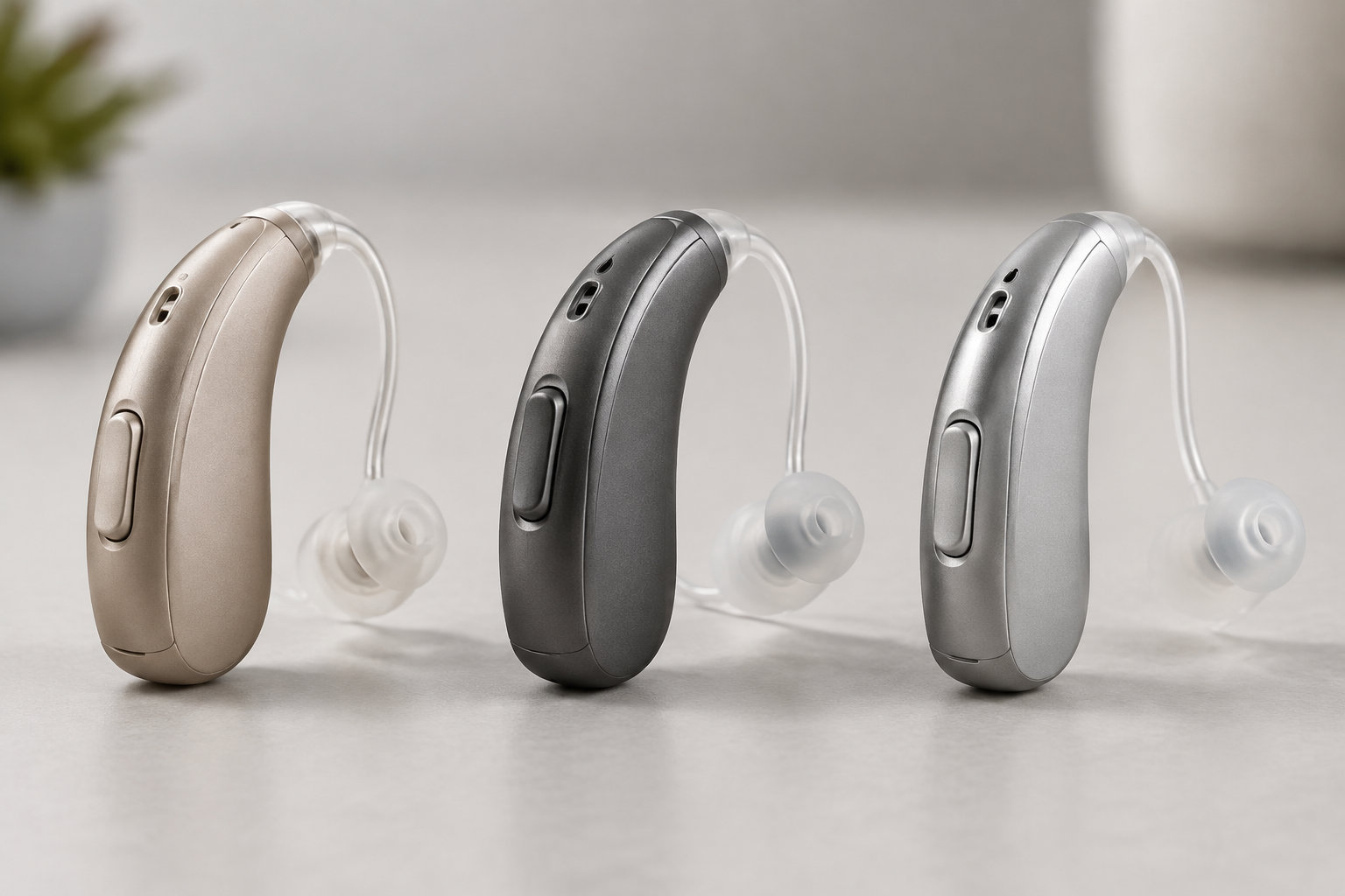 Trois appareils auditifs à contour d’oreille, de couleur beige, noire et argentée, sont alignés sur une surface claire.