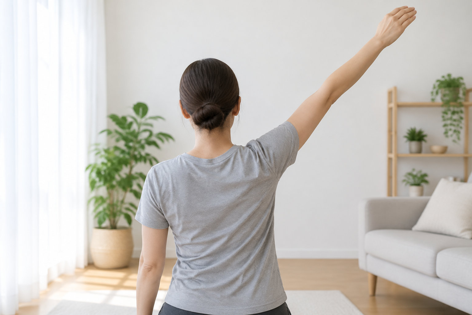 Une femme vue de dos, debout dans un salon lumineux, lève un bras en diagonale au-dessus de sa tête.