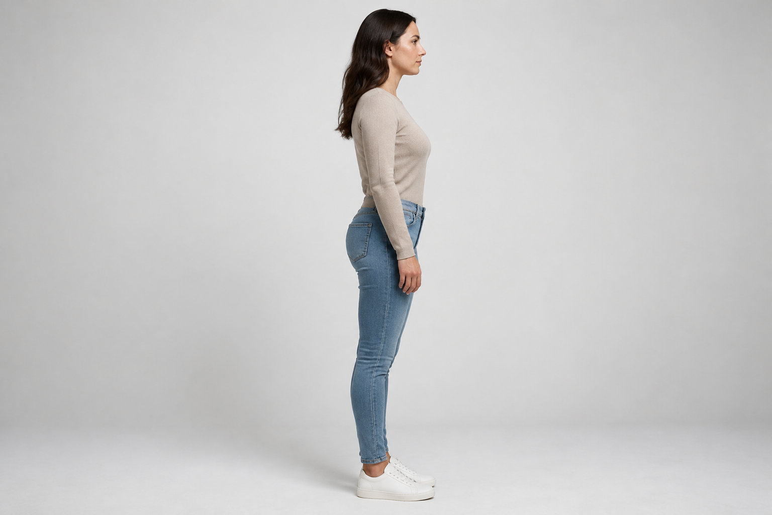 Une femme en pull beige, jean bleu et baskets blanches se tient de profil sur un fond gris clair.